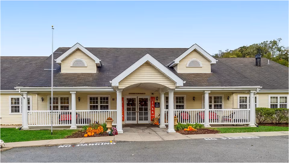Front exterior view of a single-story building with a covered entrance supported by white columns. The building has a dark shingled roof with two dormer windows and light yellow siding. There are pumpkins and fall decorations near the entrance, and a 'No Parking' sign painted on the driveway in front.