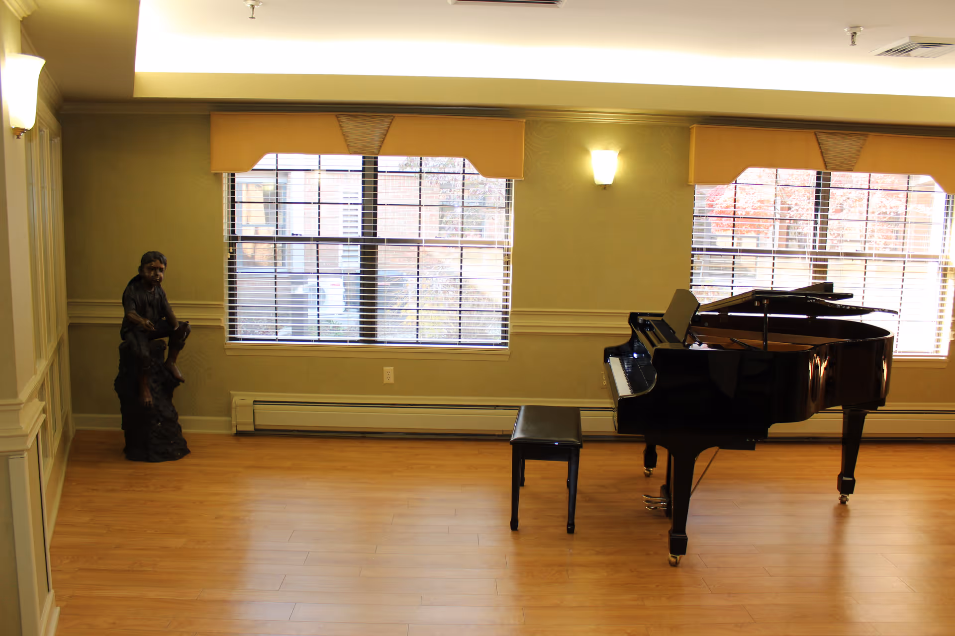 A spacious room with wooden flooring featuring a black grand piano and matching piano bench near two large windows with blinds and yellow valances. A small statue of a seated person is placed in the corner near the left window. The walls are light-colored with decorative molding and wall sconces providing soft lighting.