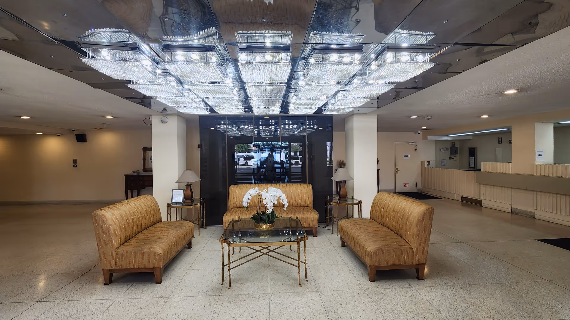 A spacious lobby area with three beige upholstered sofas arranged around a glass coffee table with a white orchid centerpiece. The ceiling features a large, reflective panel with multiple bright rectangular lights. There are two side tables with lamps beside the central sofa, and a glass entrance door is visible in the background.