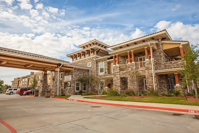 Exterior view of a large assisted living facility building with stone facade, multiple windows, and a covered entrance driveway. Several cars are parked along the driveway under a partly cloudy sky.