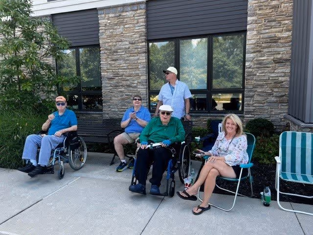 Four elderly individuals and one middle-aged woman sitting outside a building with stone and siding exterior. Two elderly men are in wheelchairs, one elderly man is sitting on a bench, and a woman is sitting on a folding chair. They appear to be enjoying a sunny day with drinks in hand.