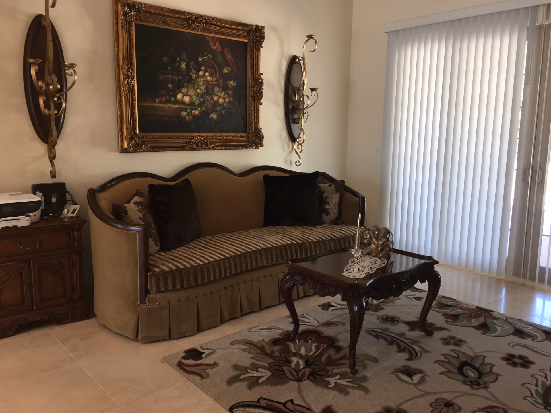 Elegant living room featuring an upholstered sofa with throw pillows, a carved wooden coffee table on a patterned rug, framed artwork on the wall, and vertical blinds over sliding doors.