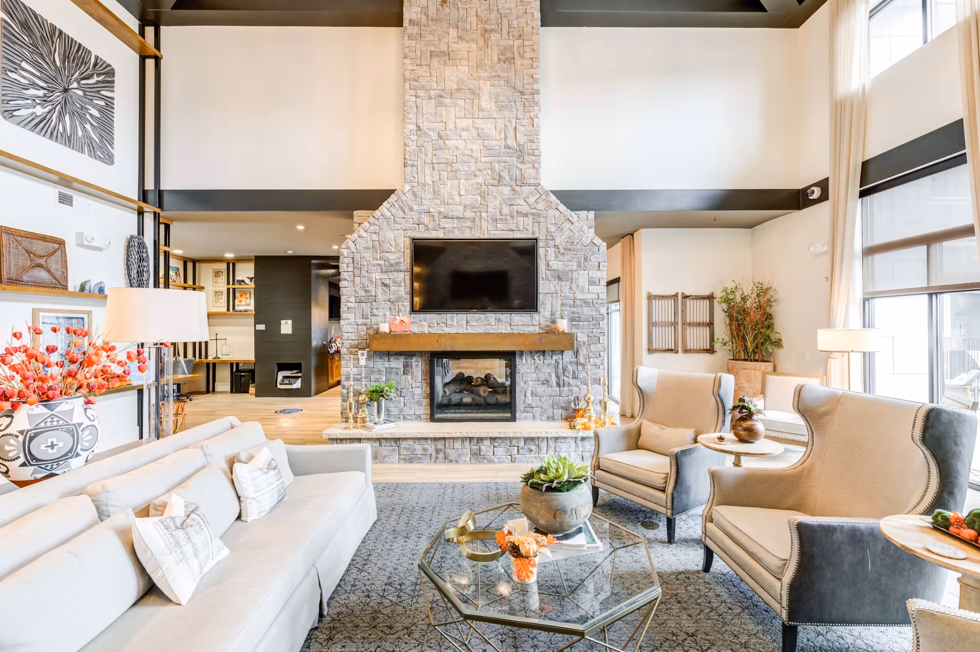 A bright and spacious living room with a large stone fireplace and a mounted flat-screen TV above it. The room features a light beige sofa with several pillows, two beige and gray armchairs with nailhead trim, and a glass coffee table with decorative items including a plant and flowers. The room has large windows with sheer curtains allowing natural light to fill the space. Decorative shelves with art pieces and a floor lamp are visible on the left side, and a potted plant is in the corner near the windows.