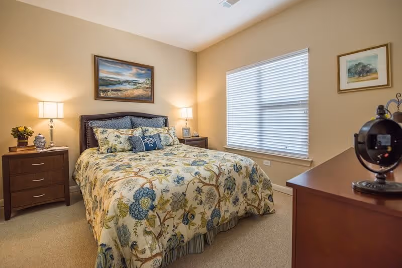 A cozy bedroom with a bed covered in a floral patterned comforter and multiple pillows. There are two wooden nightstands on either side of the bed, each with a lamp and decorative items. A large window with closed blinds is on the right wall, and two framed paintings hang on the walls. A wooden dresser with a decorative clock is visible in the foreground.