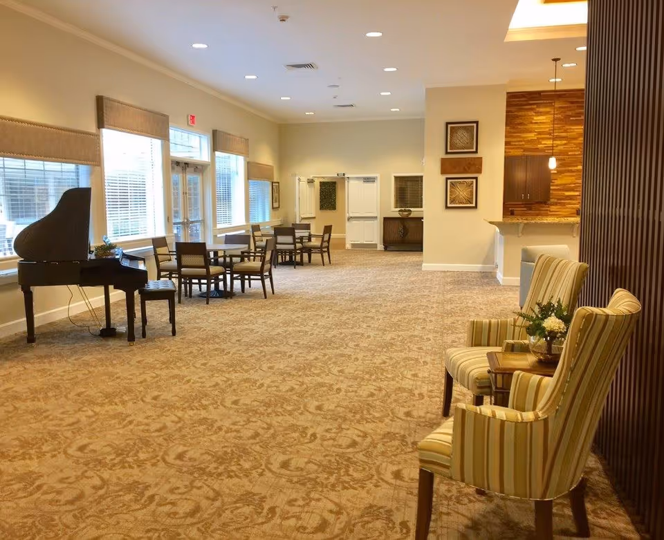 A spacious, well-lit common area with patterned carpet, several tables and chairs arranged along the windows, a black grand piano with a bench, and two striped armchairs with a small table and flower arrangement near a wooden partition. The walls are decorated with framed artwork and there is a counter with pendant lights in the background.
