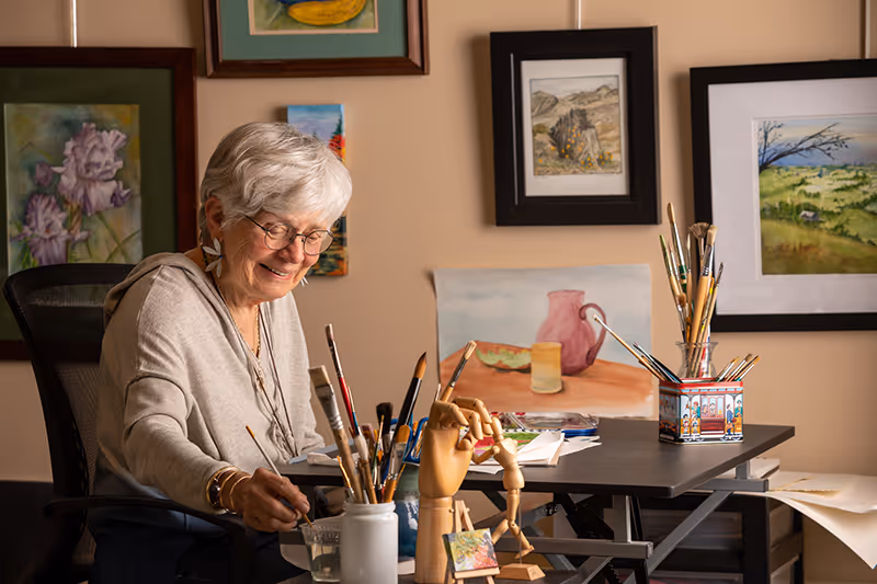 An elderly woman with short gray hair and glasses is sitting at a table painting. The table is filled with paintbrushes, art supplies, and a wooden artist's mannequin. Behind her on the wall are several framed paintings and artworks.