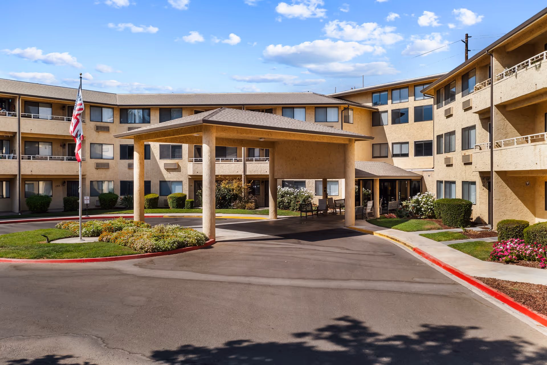 Exterior view of Walnut Park - A Provincial Senior Living Community, showing a multi-story beige building with balconies and windows. There is a covered entrance with pillars, a driveway, landscaped greenery, an American flag on a flagpole, and a clear blue sky with some clouds.