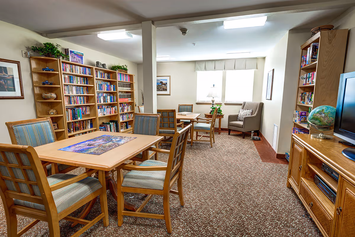 A cozy community room with bookshelves, tables and chairs, a TV cabinet, and a small seating area by the windows.