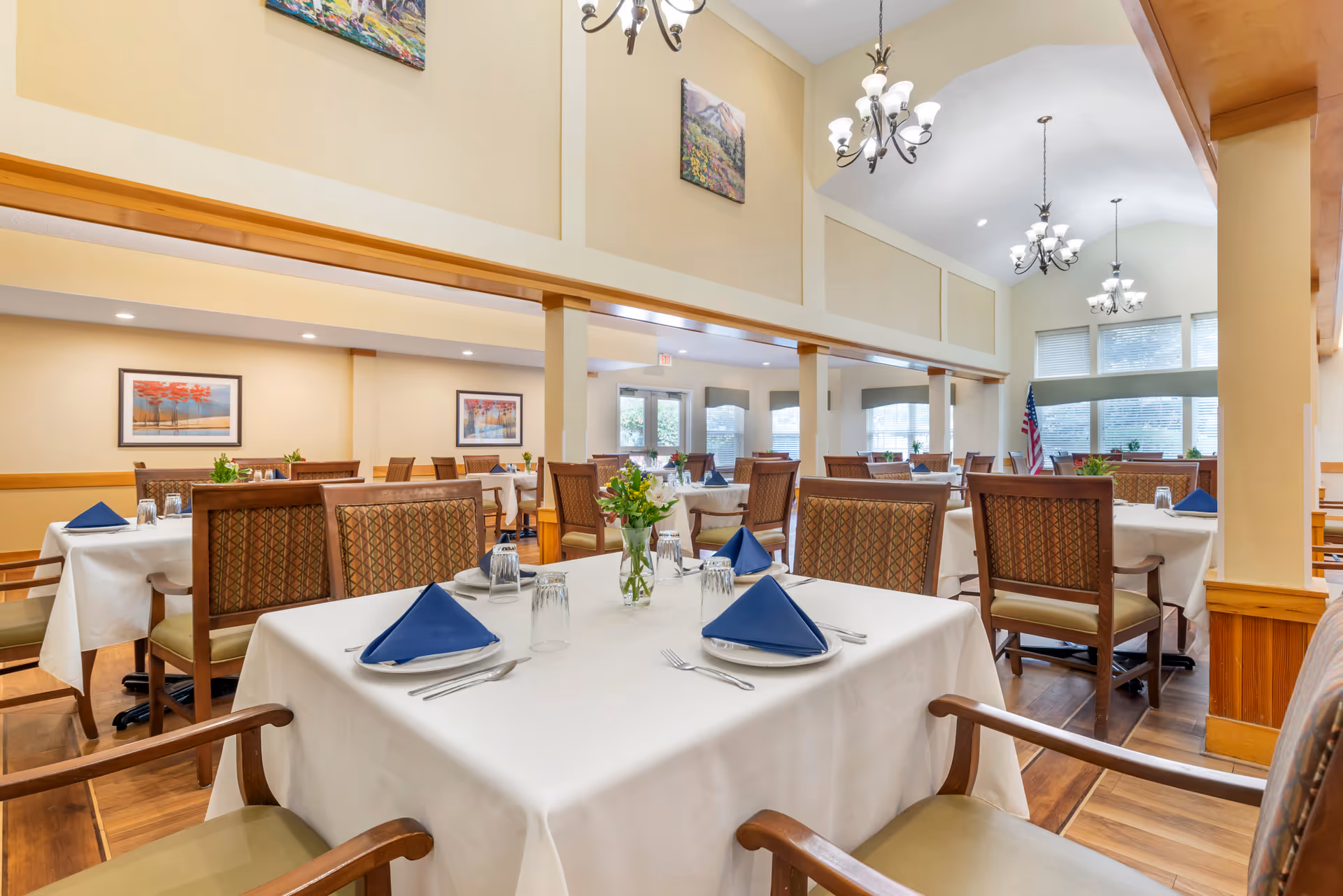 A spacious dining room with multiple tables covered in white tablecloths, each set with plates, silverware, glasses, and blue folded napkins. The room features wooden chairs with patterned upholstery, large windows letting in natural light, chandeliers hanging from a high ceiling, and framed artwork on the walls. There are small flower arrangements on each table.