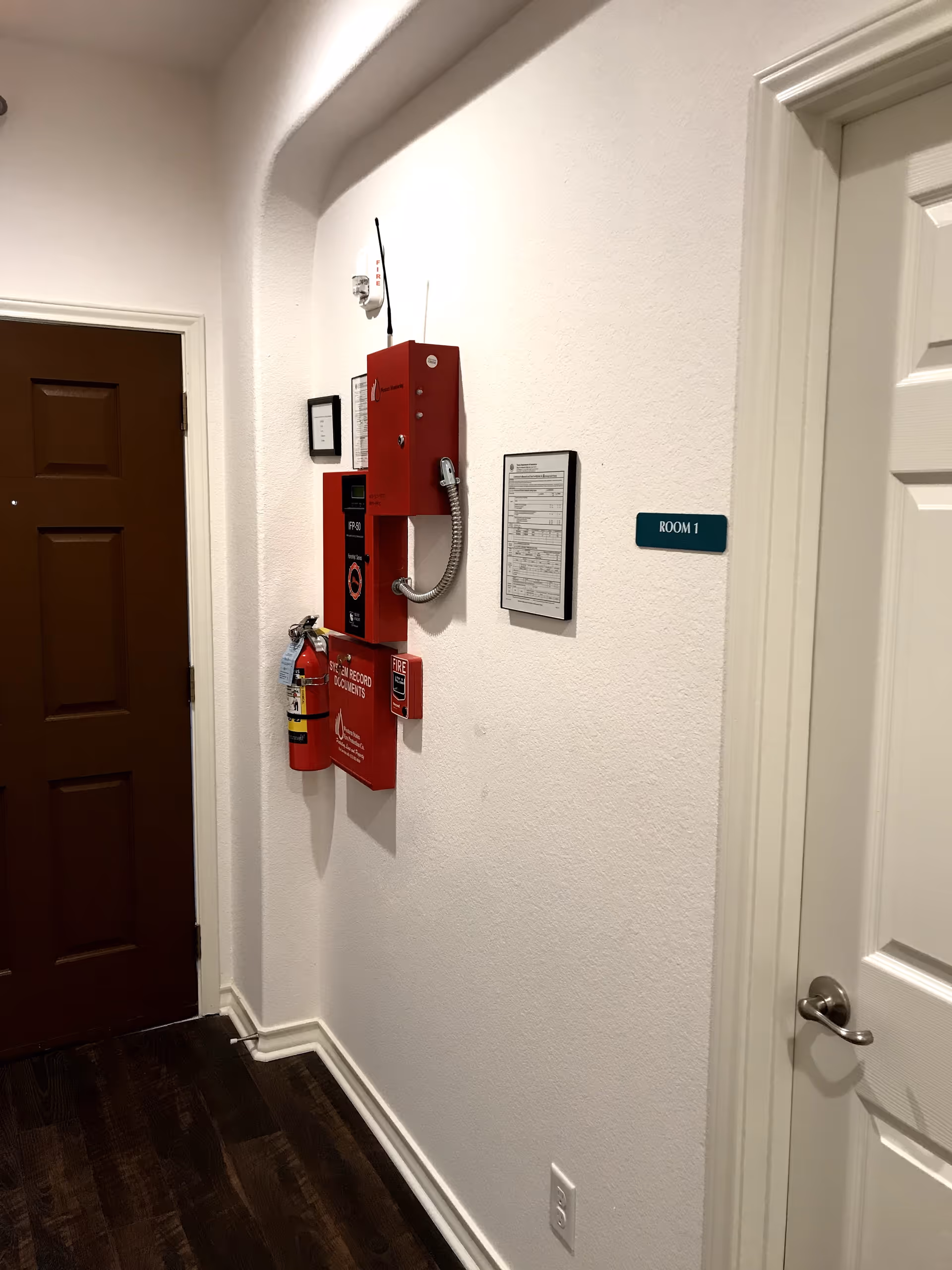 Interior hallway with white walls and dark wooden floor. On the left side, there is a brown door, and on the right side, a white door labeled 'ROOM 1'. Mounted on the wall are red fire safety equipment including a fire extinguisher, fire alarm pull station, and a fire alarm phone. Two framed documents are also visible on the wall.