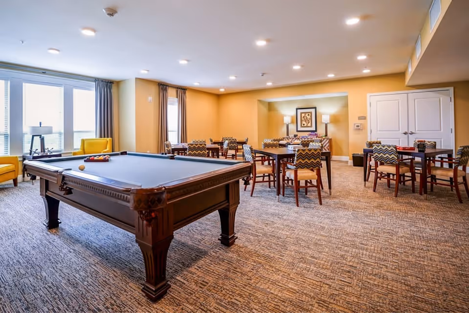 A spacious recreational room with a pool table in the foreground and several tables with chairs arranged in the background. The room has large windows with curtains, warm yellow walls, carpeted flooring, and ceiling lights. There are two yellow armchairs near the windows and framed artwork on the far wall with two lamps on a sideboard.