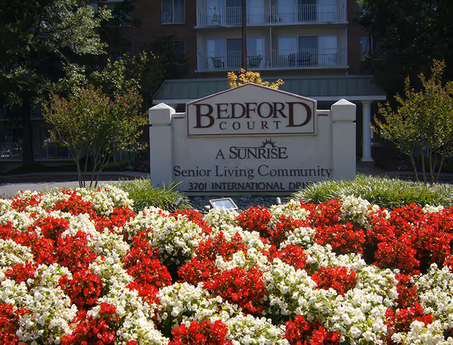 A sign for Bedford Court, a Sunrise Senior Living Community, surrounded by a garden bed filled with red and white flowers. Behind the sign, there are trees and a multi-story building with balconies.