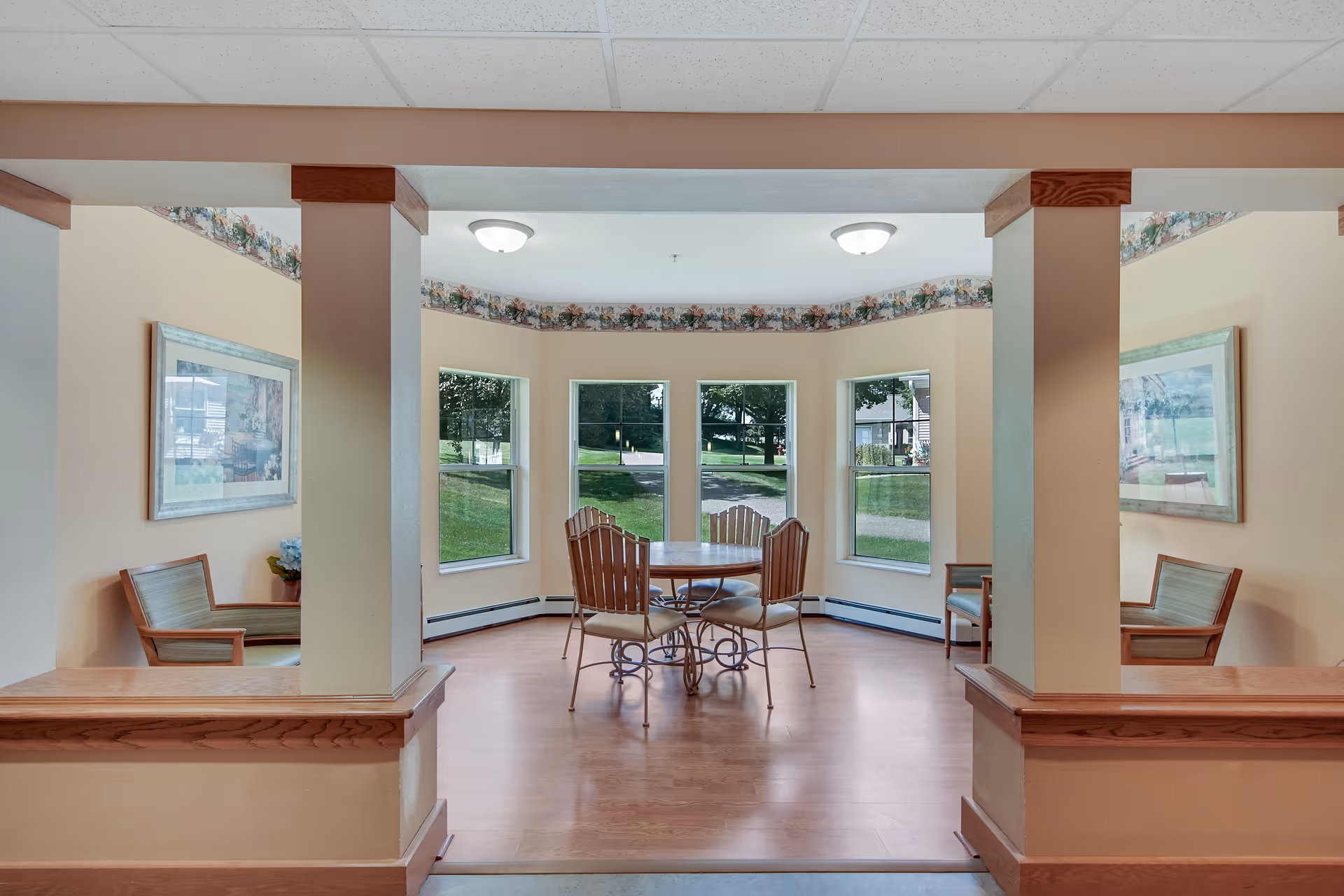 A bright communal dining area with a round table and wooden chairs centered by bay windows overlooking a lawn.