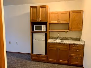 A kitchenette area featuring wooden cabinets, a small refrigerator, a microwave, and a sink, with a neutral wall and carpeted floor.