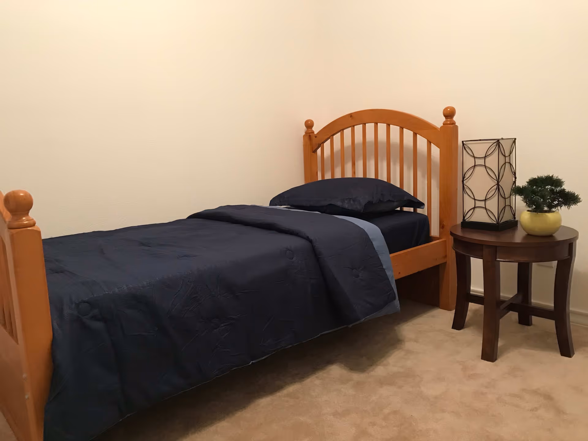 A simple bedroom with a single wooden bed featuring a dark blue bedspread and pillow. Next to the bed is a round wooden side table with a decorative lamp and a small potted plant. The room has beige carpet and plain light-colored walls.
