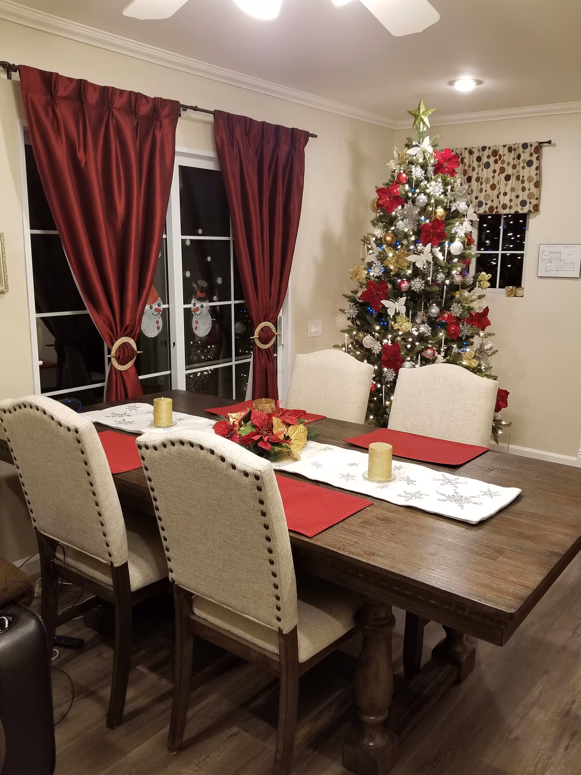 A dining room decorated for Christmas with a wooden dining table set with four beige upholstered chairs. The table has red placemats, a white table runner with snowflake designs, two candles, and a poinsettia centerpiece. In the background, there is a decorated Christmas tree with red, gold, and silver ornaments and a star on top. Red curtains frame a glass door with snowman decorations on the glass.
