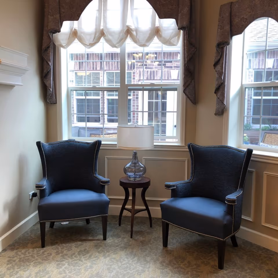 Two dark blue upholstered armchairs with nailhead trim are positioned on either side of a small round wooden table with a glass lamp on it. The seating area is in front of a large window with sheer white curtains and decorative valances. The room has beige walls and carpeted floor.