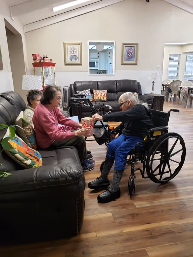 Three elderly women in a communal living room area, with one in a wheelchair reaching for popcorn from a container while others sit on sofas.