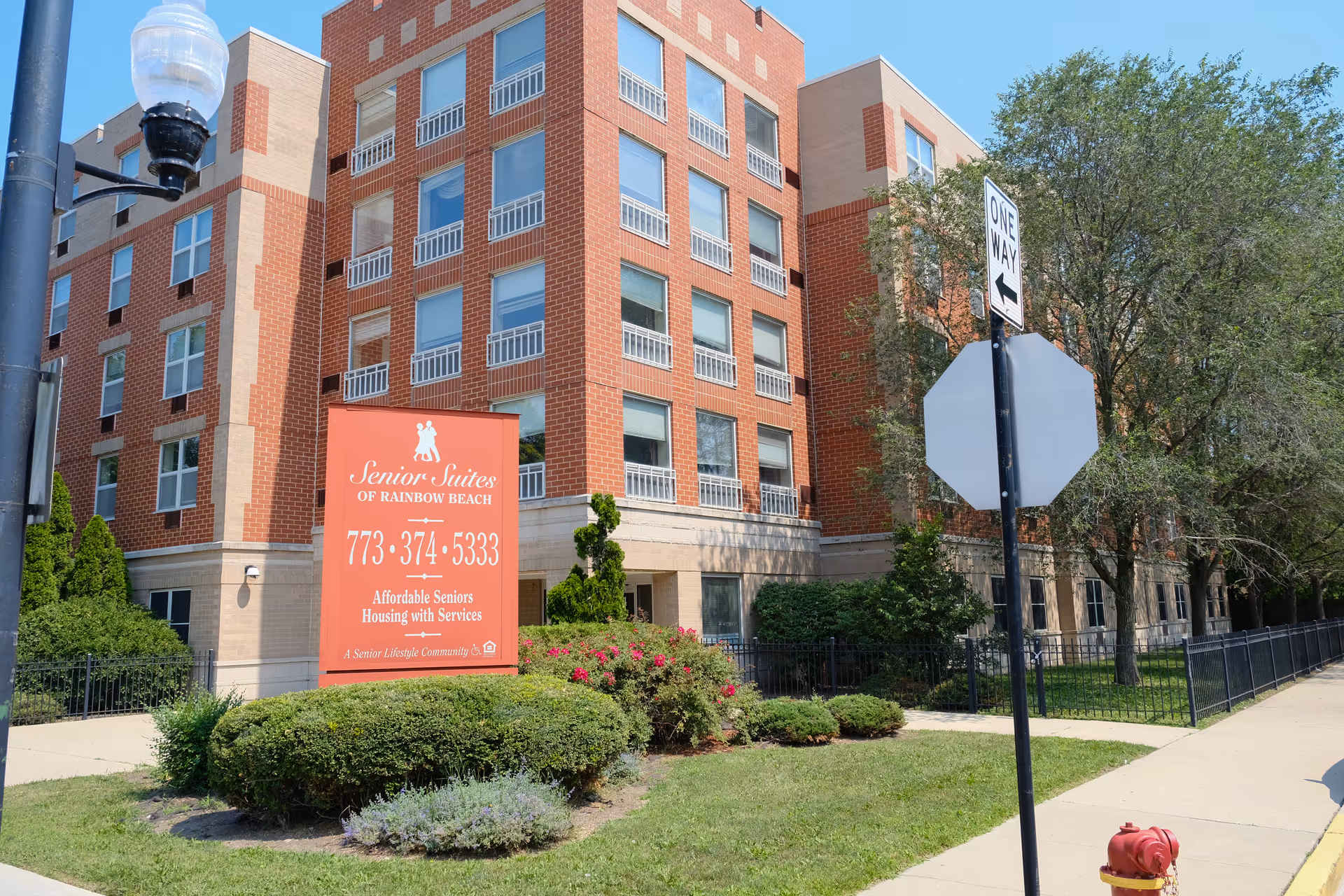 Exterior view of a multi-story brick building with multiple windows, identified as Senior Suites of Rainbow Beach. In front of the building is a large red sign displaying the facility's name, phone number, and description as affordable seniors housing with services. The area around the building is landscaped with bushes, flowers, and trees. A one-way street sign and a fire hydrant are visible on the sidewalk nearby.