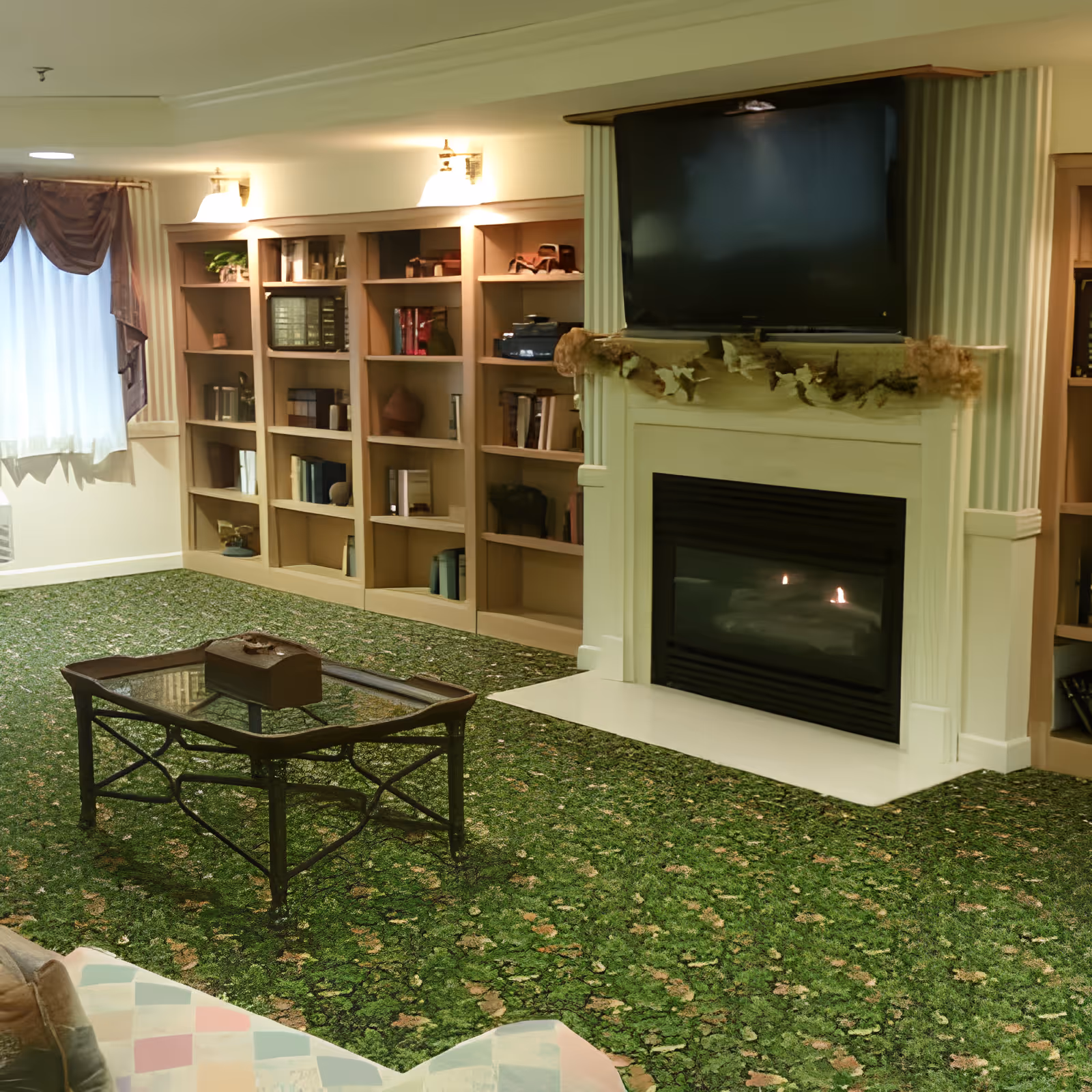 A cozy living room with a green patterned carpet, a glass coffee table with a wooden box on top, built-in bookshelves filled with books and decorative items, a fireplace with a TV mounted above it, and a window with curtains allowing natural light in.