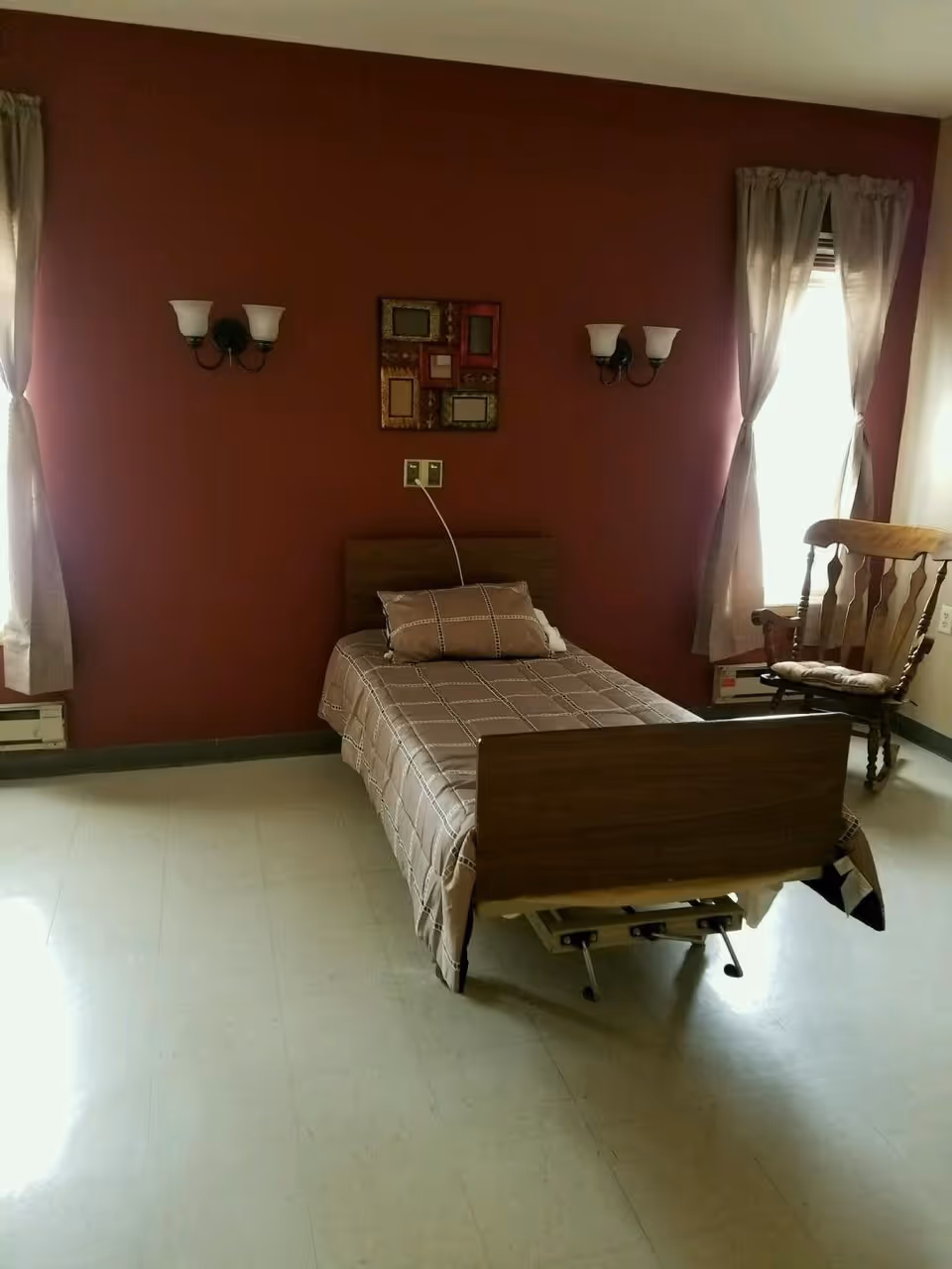 A simple bedroom with a single bed covered in a brown checkered bedspread and a matching pillow. The bed has a wooden headboard and footboard. The room has two windows with beige curtains, a wooden rocking chair with a cushion near one window, and two wall-mounted light fixtures with frosted glass shades on a dark red wall. A decorative multi-frame wall art hangs above the bed.