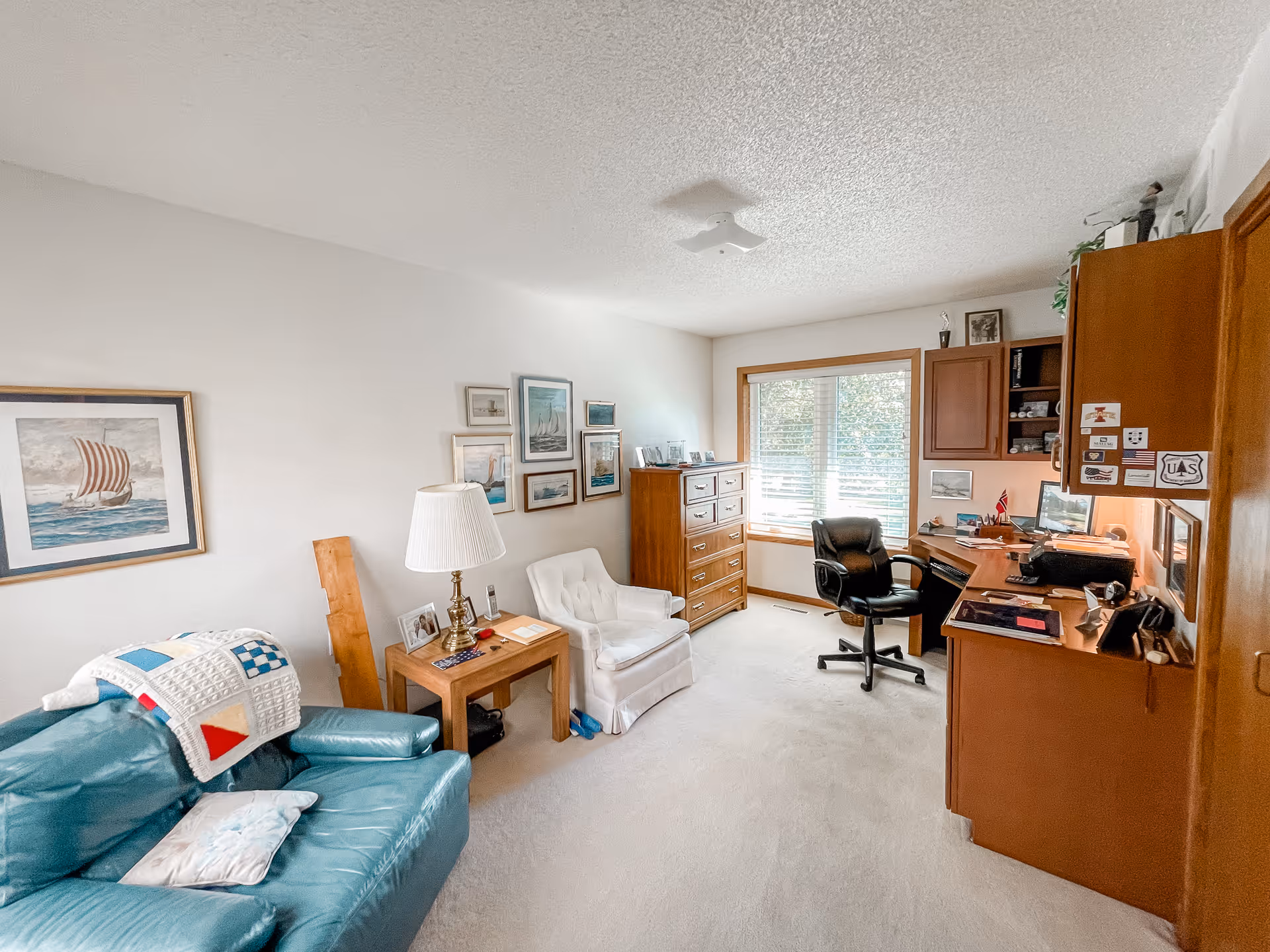 A cozy room with a teal leather couch, a white armchair, a wooden side table with a lamp, and a wooden dresser. The walls are decorated with framed pictures, mostly of boats and nautical themes. There is a wooden desk with a black office chair near a window with white blinds, and wooden cabinets above the desk. The room has light-colored carpet and a textured ceiling.