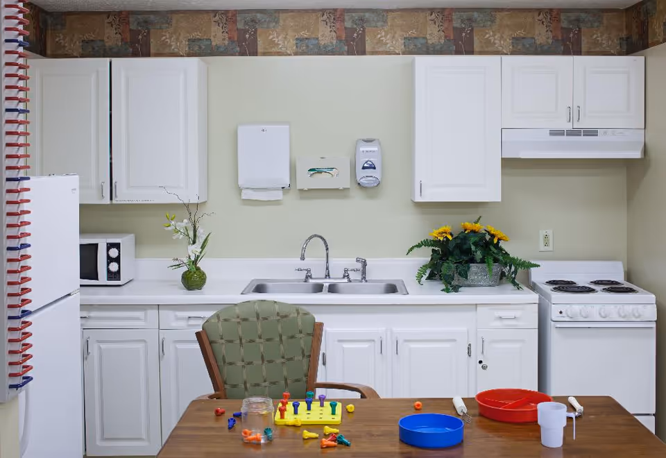 A small kitchen area with white cabinets, a double sink, a white stove, and a white refrigerator. On the countertop, there is a microwave, a green vase with white flowers, and a planter with yellow flowers. A wooden table with a green cushioned chair is in front of the sink, with colorful game pieces, a red divided plate, a blue plate, and a white cup on the table. The wall above the cabinets has a decorative border.