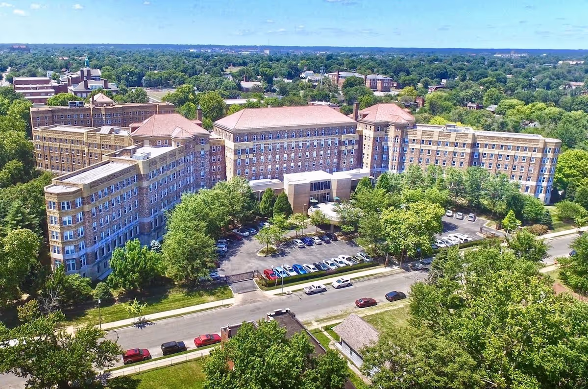 Aerial view of a large multi-story brick building complex surrounded by trees and greenery, with a parking lot filled with cars in front. The building is situated in a suburban area with more buildings and trees visible in the background under a clear blue sky.