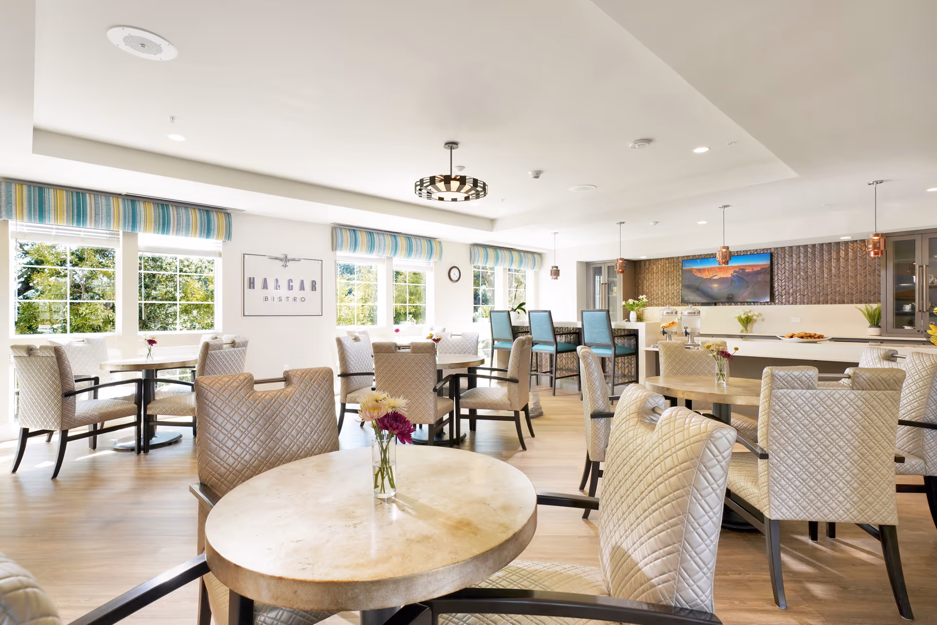 Bright and spacious dining room with round tables and cushioned chairs, large windows with striped valances letting in natural light, a bar area with high chairs, and a wall-mounted TV above a counter with decorative plants and refreshments.