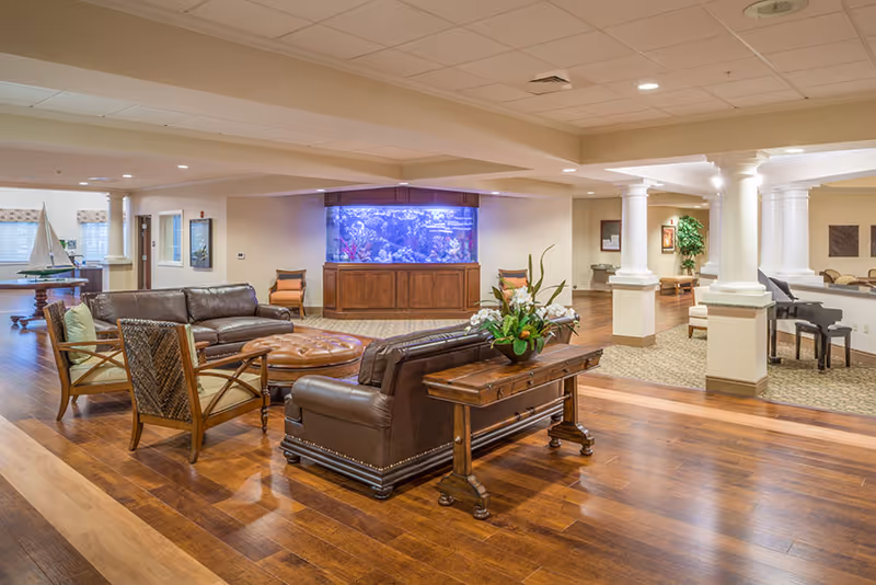 Spacious assisted living facility common area with wooden flooring, leather sofas, wooden chairs, a large aquarium built into the wall, and a piano in the background. The room is well-lit with ceiling lights and decorated with plants and artwork.