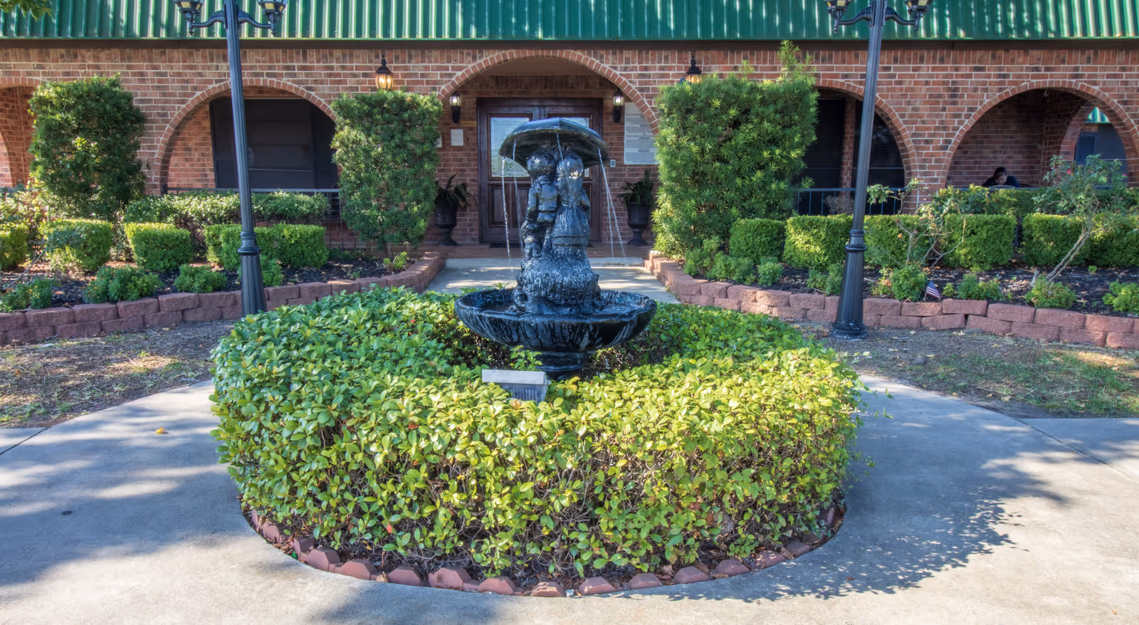 Front entrance of a brick building with a central decorative fountain sculpture surrounded by trimmed hedges and walkways.