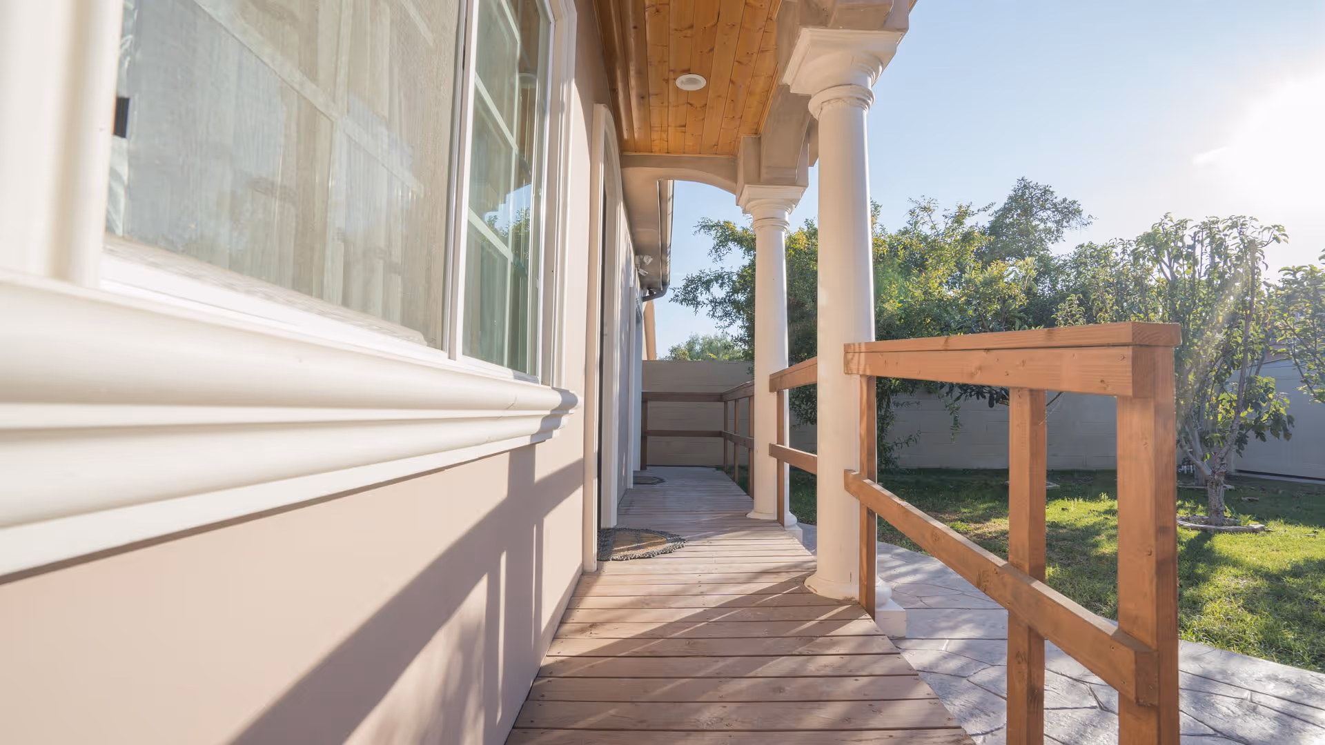 A wooden porch with white columns and a wooden railing overlooking a green yard with trees under a clear sky.