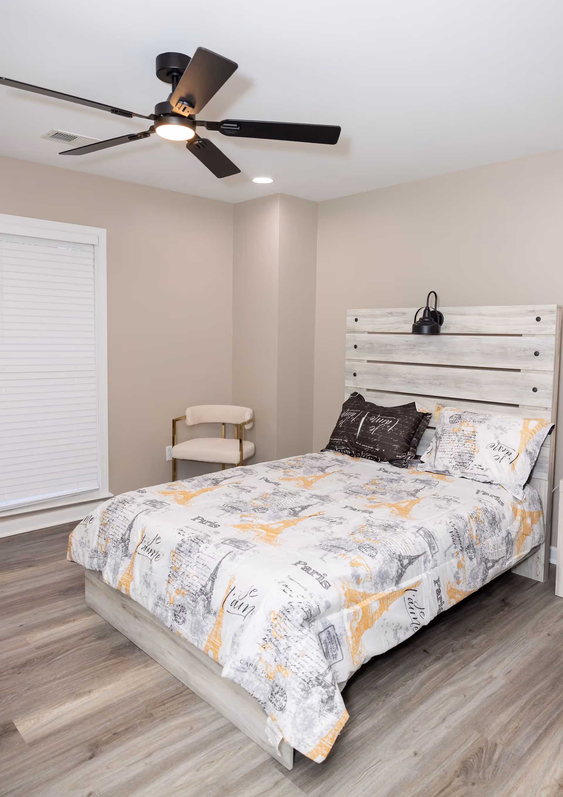 A bedroom with a bed featuring a light wood headboard and bedding decorated with Eiffel Tower and Paris-themed prints. There is a beige upholstered chair with gold metal arms in the corner, a ceiling fan with black blades and a light, and a window with closed white blinds. The floor is light wood, and the walls are painted beige.