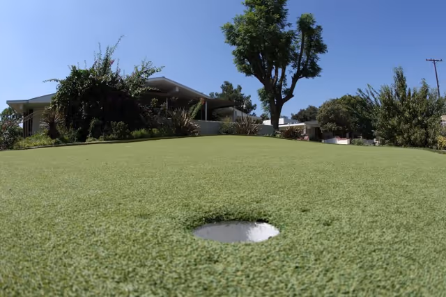 A putting green with a golf hole in the foreground, surrounded by bushes and trees, with a building partially visible in the background under a clear blue sky.
