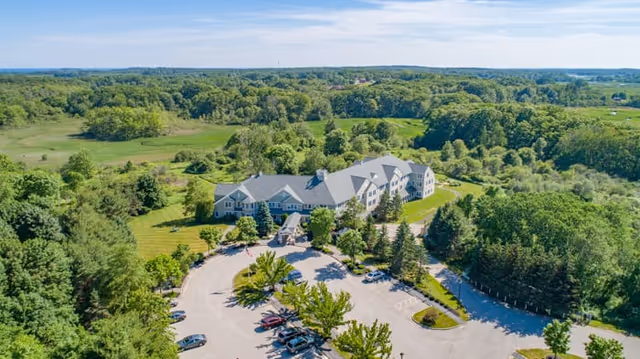 Aerial view of The Landing at Cape Elizabeth facility surrounded by lush green trees and open fields, with a large parking lot in front of the building under a clear blue sky.
