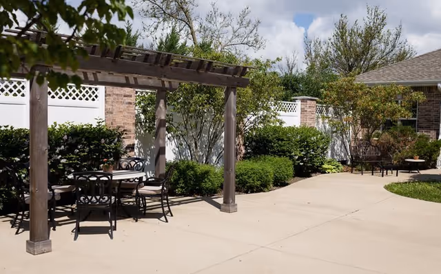 Outdoor patio area with a wooden pergola covering a round table and chairs. The space is surrounded by green bushes, trees, and a white fence, with a brick building partially visible in the background under a partly cloudy sky.