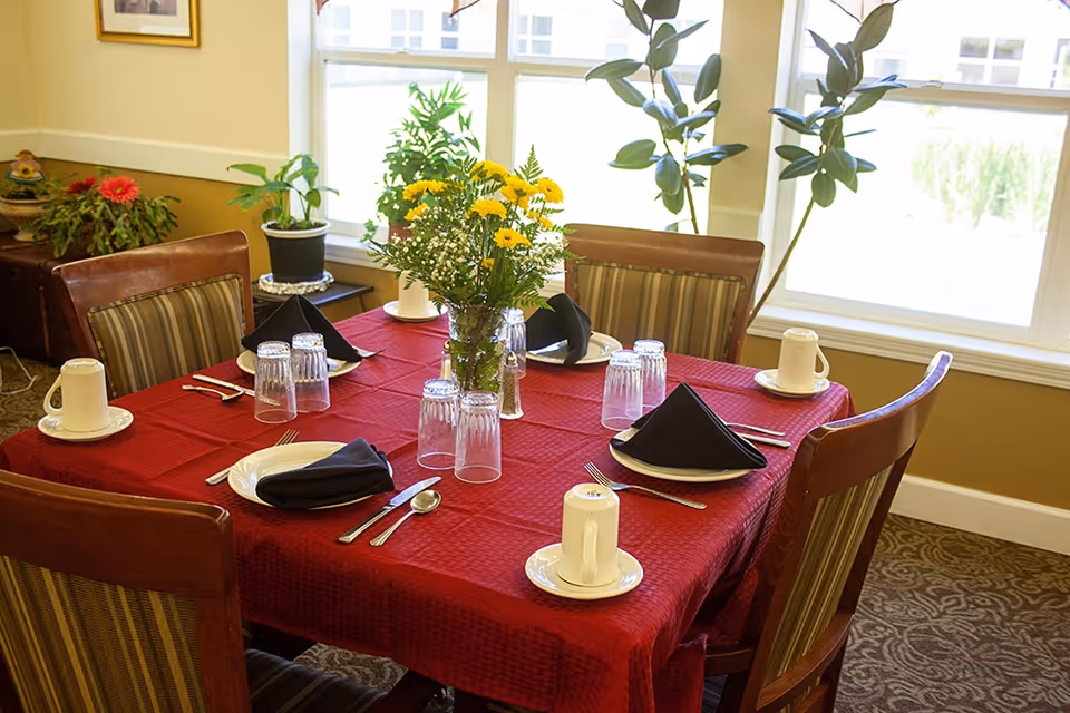 A dining table set for four with a red tablecloth, white plates, black folded napkins, upside-down drinking glasses, and white coffee cups on saucers. A vase with yellow and white flowers is in the center of the table. The table is surrounded by four wooden chairs with striped cushions. There are large windows in the background letting in natural light, and some potted plants are visible near the windows.