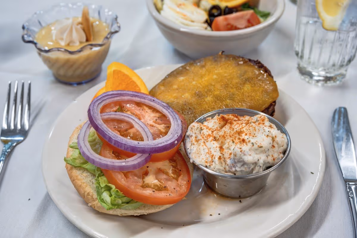 A plate with an open cheeseburger featuring lettuce, tomato slices, and red onion rings on the bottom bun, and a melted slice of cheddar cheese on the top bun. Next to the burger is a small metal cup filled with a creamy salad topped with paprika. In the background, there is a bowl of salad and a glass of water with a lemon slice. A fork and knife are placed on either side of the plate on a white tablecloth.