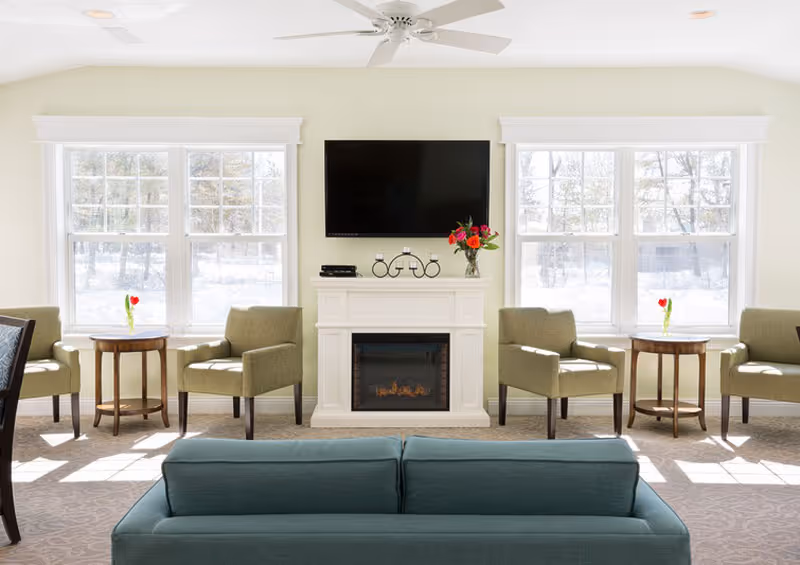 Bright communal living room with a sofa facing a fireplace and wall-mounted TV, flanked by armchairs and large windows.