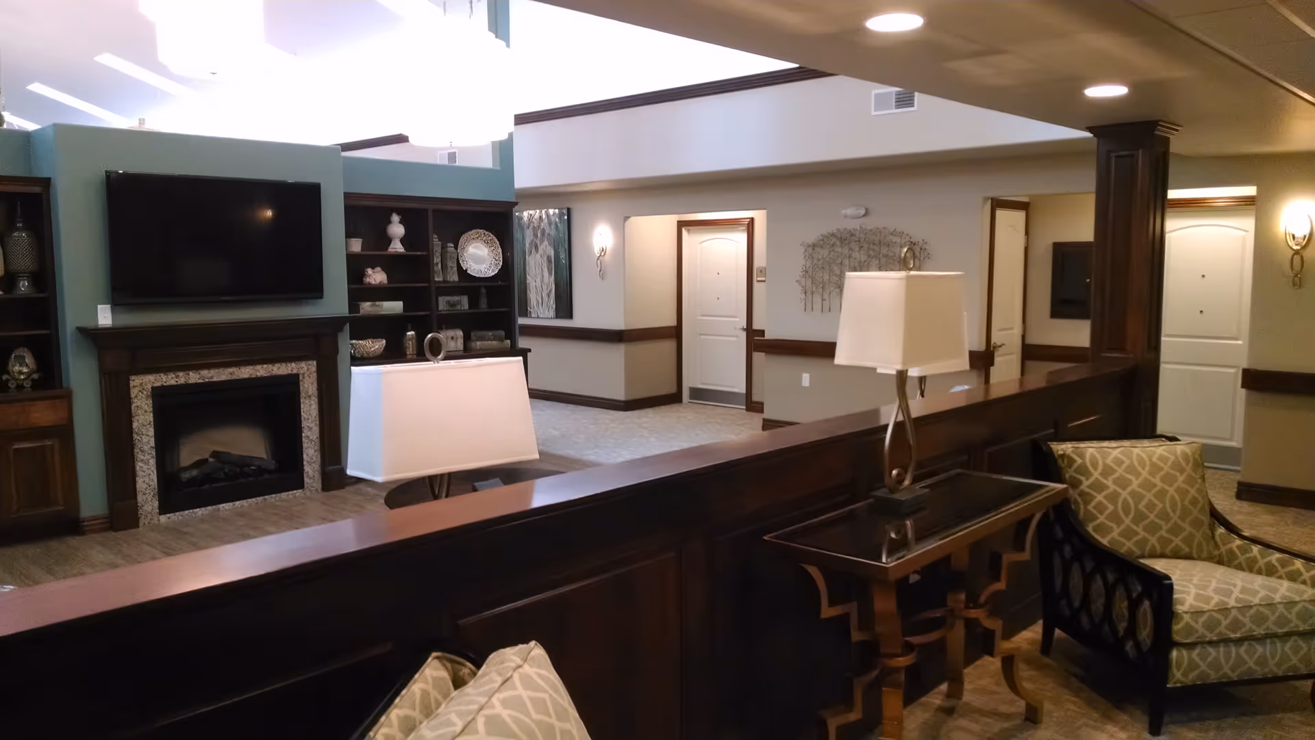 Interior view of a senior living facility common area featuring a wall-mounted flat-screen TV above a fireplace, built-in shelves with decorative items, a wooden partition with table lamps, and cushioned armchairs. The space has beige walls, carpeted floors, and multiple doors leading to other rooms.