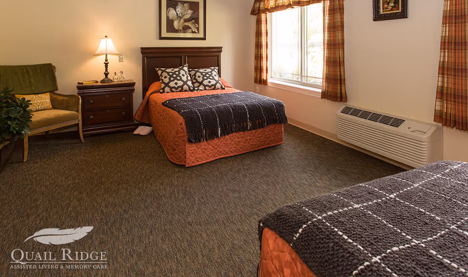 A cozy bedroom in an assisted living facility featuring two beds with orange bedspreads and dark gray knitted throws, a wooden nightstand with a lamp, a green armchair with a cushion, a large window with plaid curtains, and a wall-mounted air conditioning unit.