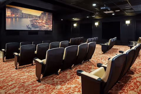Interior view of a small movie theater room with multiple rows of black and beige cushioned seats facing a large screen displaying an image of a canal cityscape at sunset. The room has patterned carpet flooring and wall sconces providing soft lighting.