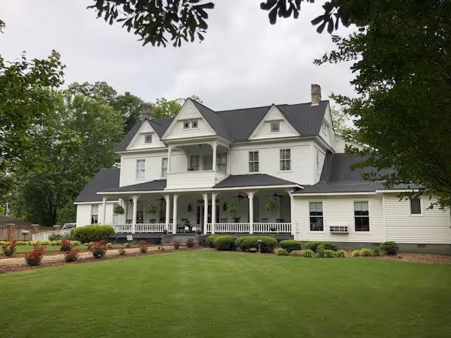 Large white two-story house with a black roof, a spacious front porch with hanging plants, surrounded by green lawn and trees under a cloudy sky.