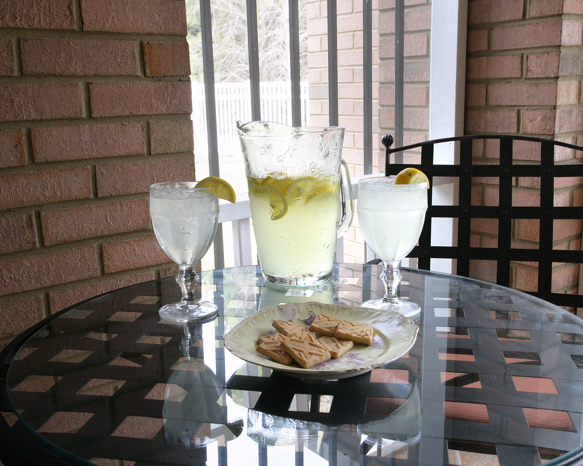 A glass table on a patio with a pitcher of lemonade containing lemon slices, two glasses of water with lemon wedges on the rims, and a plate with several rectangular cookies. The background shows brick walls and metal railing.
