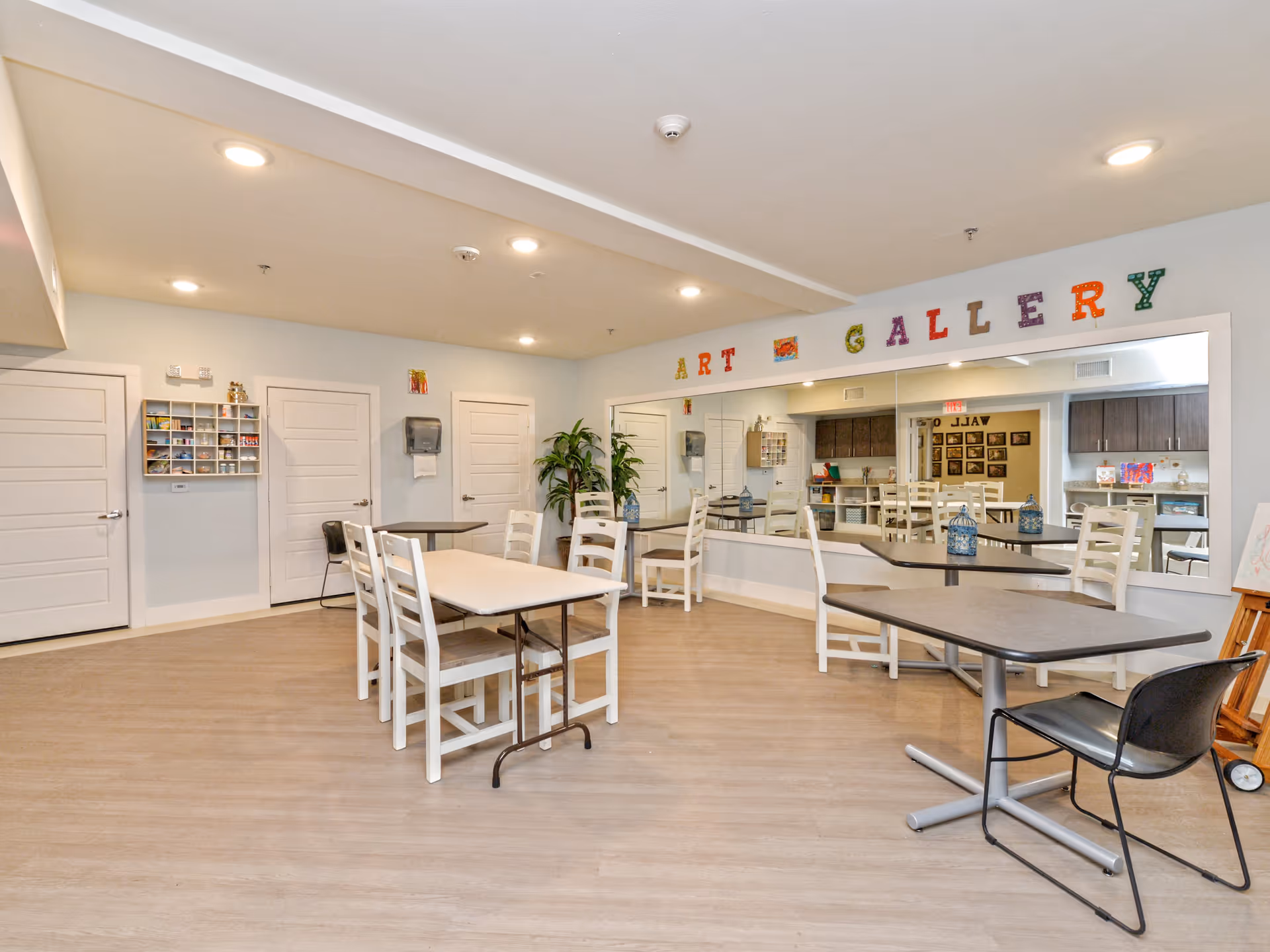A bright and spacious room with light wood flooring and white walls, featuring several tables and chairs arranged for group activities. On the far wall, colorful letters spell out 'ART GALLERY' above a large mirror. There are three closed white doors, a wall-mounted paper towel dispenser, a small shelving unit with art supplies, and a potted plant in the corner. The room appears to be designed for creative or social activities in a senior living facility.