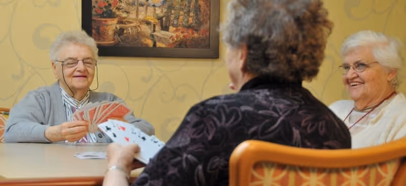 Three elderly women sitting around a table playing cards in a room with yellow patterned wallpaper and a framed painting on the wall.