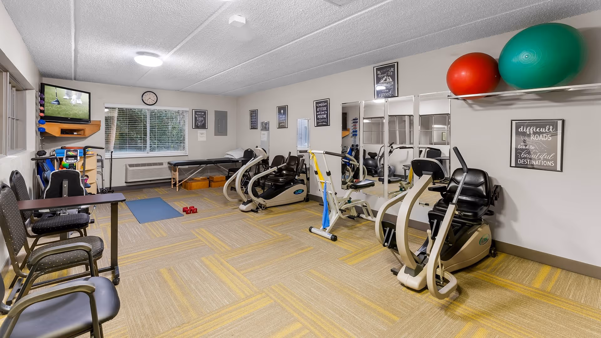 A fitness room in a senior living facility with exercise equipment including stationary bikes, a recumbent bike, a massage table, exercise balls on a wall shelf, a yoga mat, and small dumbbells. There are chairs along one wall, a TV mounted in the corner, and motivational posters on the walls. A window with blinds lets in natural light.