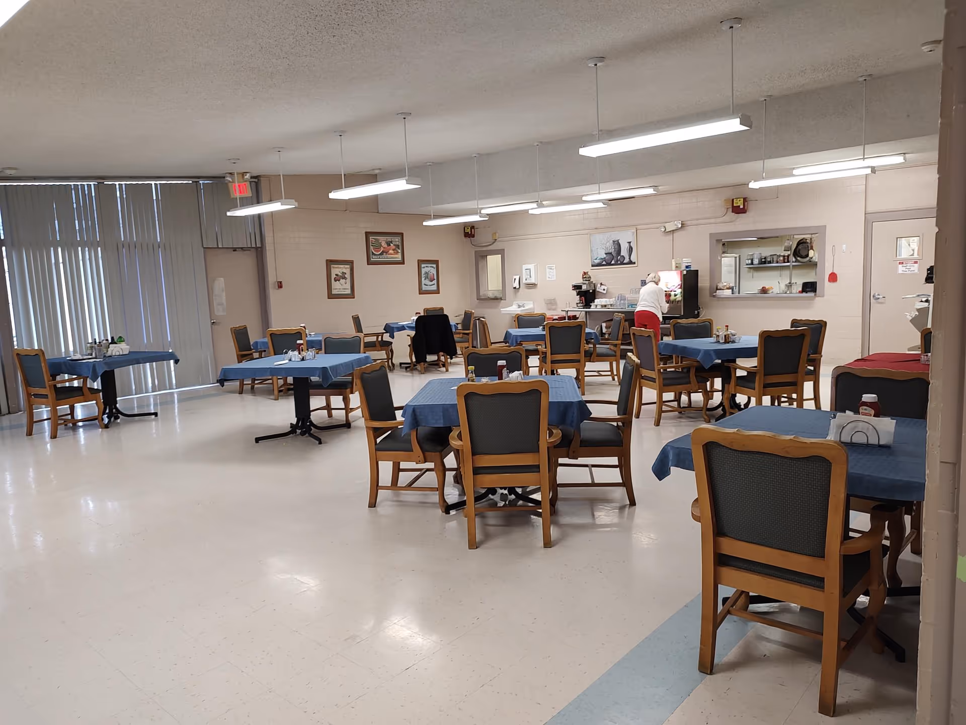 Spacious dining room with multiple tables covered in blue tablecloths, wooden chairs, and a staff member at the service counter.