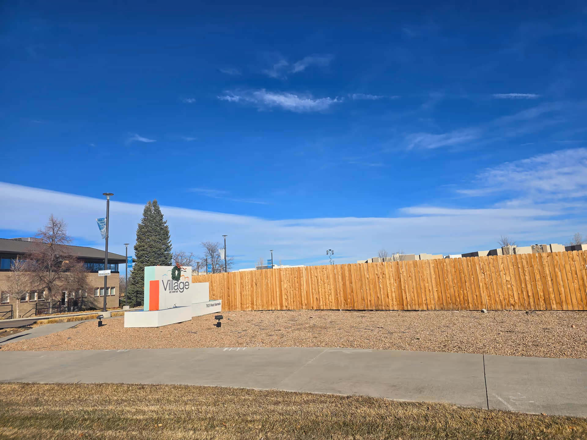 Outdoor view of the Village at Belmar facility sign with a wooden fence and building in the background under a clear blue sky.