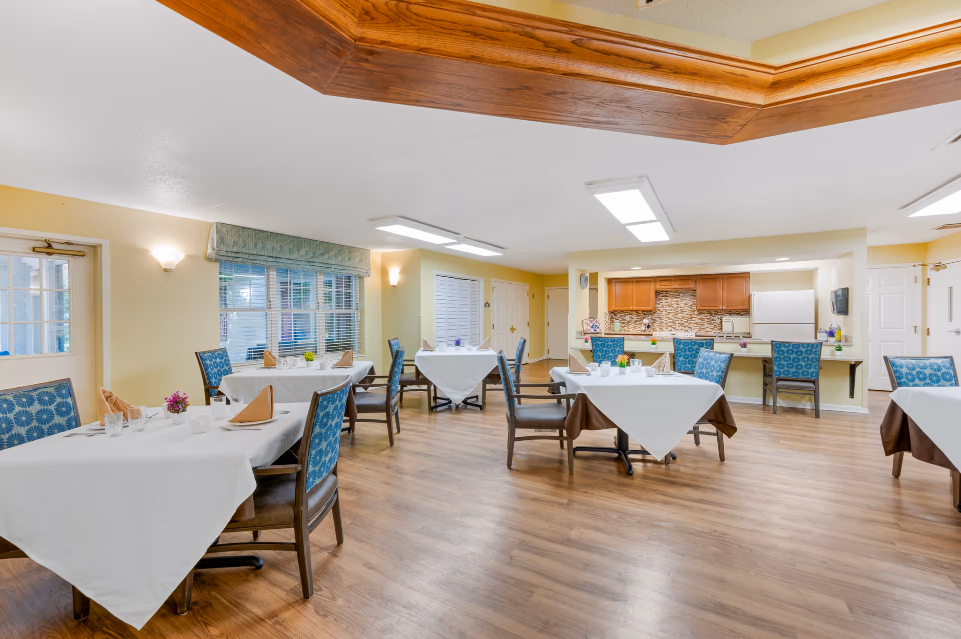 A bright dining room with several tables covered in white tablecloths and brown underlays, each set with napkins, glasses, and small flower arrangements. The room has wooden flooring, blue patterned chairs, and a kitchen area with wooden cabinets and a white refrigerator in the background. There are windows with blinds and a door on the left side, and the ceiling features recessed lighting and a wooden trim detail.